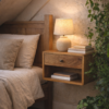 Square floating bedside table made from solid mango wood in an oak finish, wall-mounted beside an oak bed in a cosy attic bedroom, styled with a ceramic lamp, stacked books and trailing green plants.