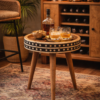 Round mango wood side table with black and white bone inlay detailing, styled in a home bar setting on an Indian-patterned rug, topped with whisky glasses, a bottle, and bar accessories.