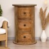 Round solid wood three-drawer bedside cabinet in a warm natural finish, styled in a cozy room with pampas grass, a woven chair, and soft lighting.