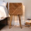 Wooden line-carved two-drawer side table with Scandinavian legs, styled beside a bed in warm natural light with a stack of books on the floor.
