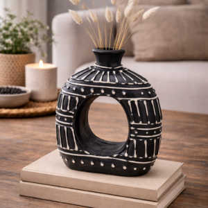 Black donut shaped ceramic vase with white tribal pattern, styled on stacked books with dried grasses on a wooden coffee table in a neutral living room setting