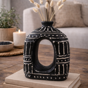 Black donut shaped ceramic vase with textured white detailing, styled on stacked books with dried grasses on a wooden table in a warm neutral living room setting