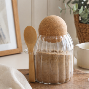 Glass storage jar with cork-style lid and wooden spoon, filled with brown sugar, styled on a rustic wooden board in a softly lit kitchen setting with neutral décor.