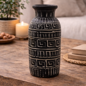Black patterned ceramic decorative vase with carved geometric detailing, styled on a wooden table with books and greenery in a warm, modern living room setting.