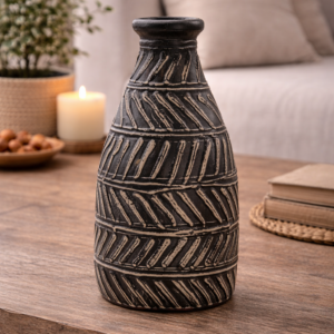 Black textured ceramic decorative vase with carved diagonal pattern, styled on a wooden table with books and greenery in a warm, neutral living room setting.