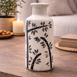 Cream ceramic decorative vase with black bamboo pattern, styled on a wooden coffee table with books, a candle and greenery in a warm, neutral living room setting.