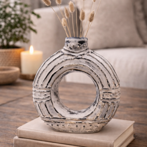 Textured circular ceramic decorative vase with distressed cream finish, styled on stacked books on a wooden coffee table with dried stems, candle and greenery in a warm, neutral living room setting.