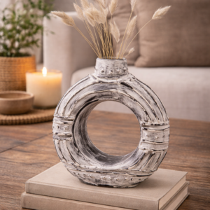 Cream donut-shaped ceramic vase styled on a wooden coffee table with dried grasses, stacked books, and soft neutral living room décor in the background