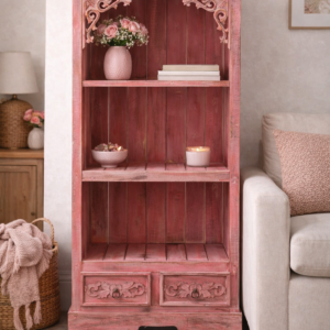 Pink wooden open cabinet with carved detailing and two drawers, styled in a neutral living room with soft furnishings, decorative accessories, and warm natural light.