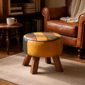 Patchwork leather footstool with wooden legs placed in a cosy reading corner beside a brown leather armchair, bookshelf and wooden side table with a coffee cup.
