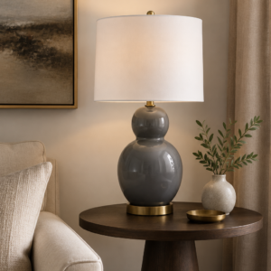 Grey ceramic table lamp with rounded base and white fabric shade, styled on a bedside table in a modern neutral bedroom with soft ambient lighting.