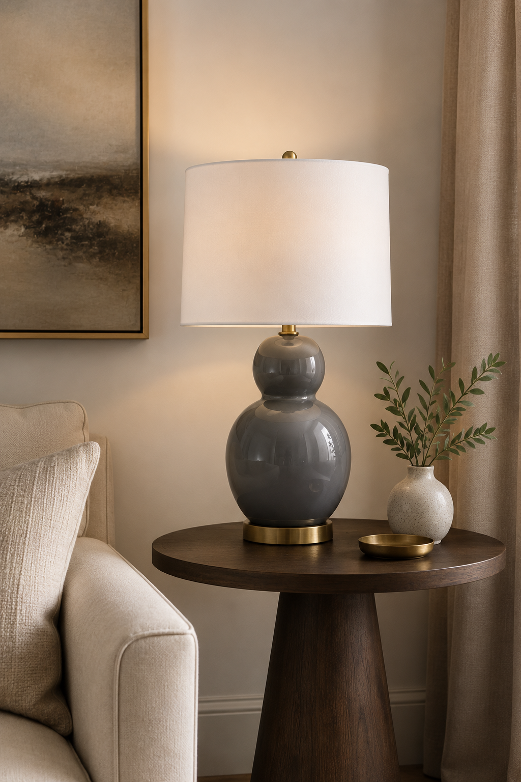 Grey ceramic table lamp with rounded base and white fabric shade, styled on a bedside table in a modern neutral bedroom with soft ambient lighting.